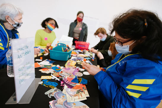 5 people of all ages seated and standing around a table covered in art making materials.