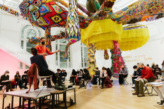 Visitors in the Paine Gallery sitting under a large colorful hanging sculpture drawing from two live models posed on a stage.