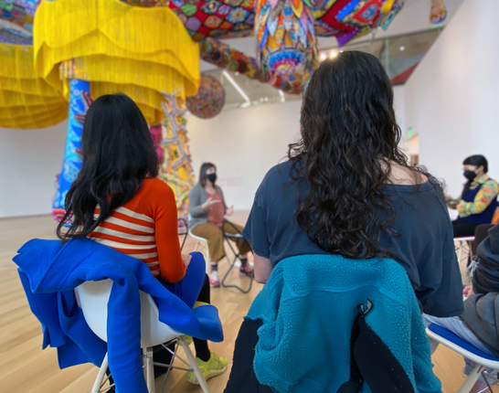 Visitors sit in a circle under a colorful sculpture and installation.