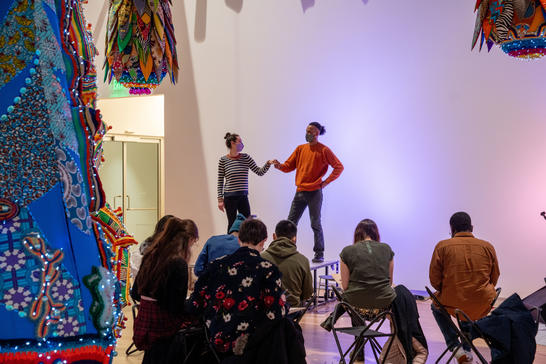 Two clothed figure models pose on a stage while visitors sketch under Joana Vasconcelos' sculpture Valkyrie Mumbet