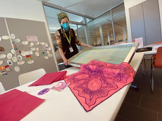 An artist holds a silkscreen with brightly colored and decorated bandanas in on a table