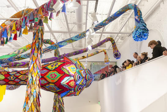 Viewers stand on a balcony in the museum looking out at the brightly colored huge sculpture hanging from the ceiling