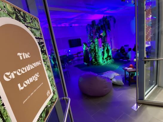 An open doorway with a sign that reads "The Greenhouse Lounge" leading into a room lit with deep purple and green lights and has low seating 