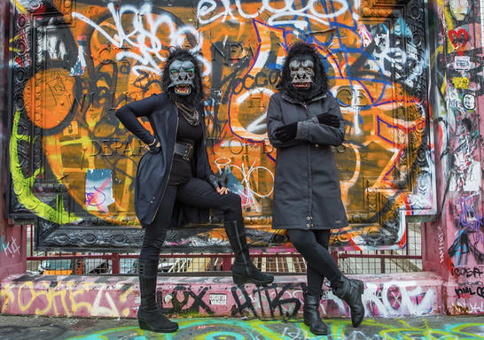Two women dress in all black with guerrilla masks stand in front of a wall covered with grafitti 