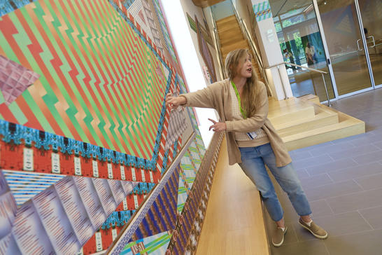 MAAM Educator with long blonde hair wearing a tan cardigan with blue jeans with her arms outstretch giving a talk about artwork on the wall 