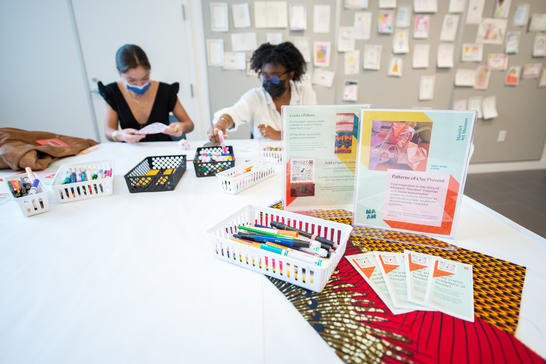 Two masked people sitting at a table with markers and cards.