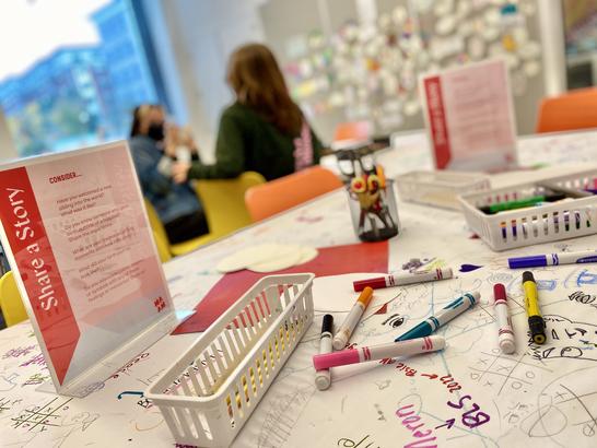 A table with markers and paper with two people in the background