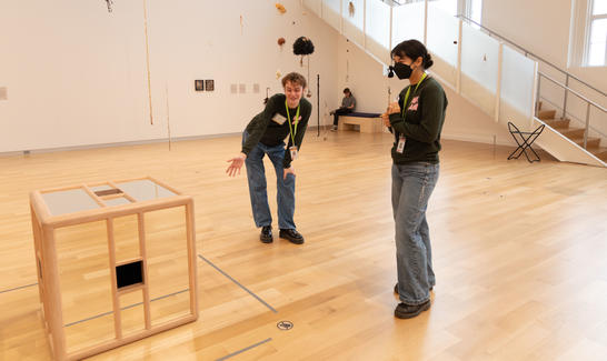 One person standing with a mask beside another person pointing at a mirrored cube.