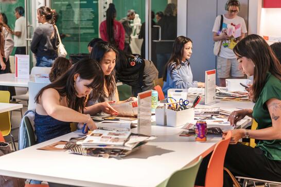 A group of people sitting at tables, engaged in arts and crafts with magazines and scissors. The setting is lively, with colorful chairs and a creative atmosphere.