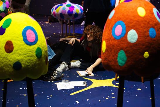 A person with curly hair sits on a starry floor, surrounded by colorful, spotted sculptures resembling mushrooms. They are drawing on a clipboard, creating a thoughtful ambiance.