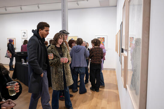 A lively art gallery scene with people admiring framed artworks on white walls. Visitors, casually dressed, hold drinks, showing engagement and interest.