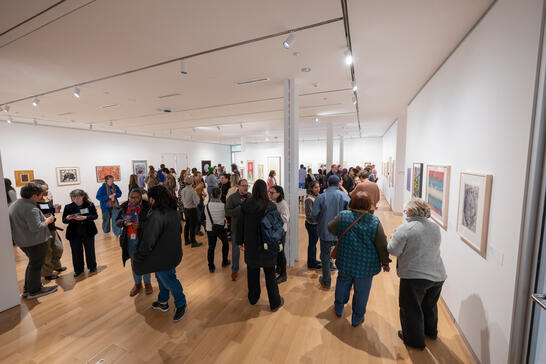 A crowded art gallery features diverse visitors engaging with colorful artworks on white walls. The scene conveys a lively, social atmosphere.