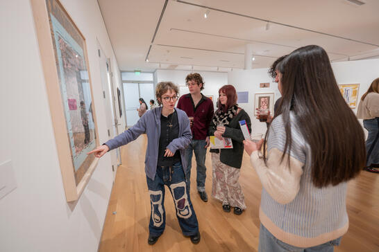 A group of people engage in discussion at an art gallery, focused on a framed piece. The setting is bright with wooden floors and white walls.
