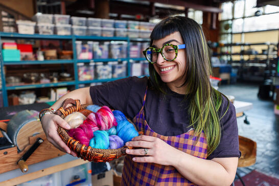 Alex Makes Art (Alex Adamo) with glasses smiles warmly in a colorful studio, holding a basket of vibrant wool yarns. Shelves with supplies fill the background.
