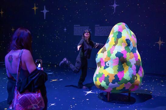 A person poses playfully beside a colorful, abstract sculpture in a dark, starry-themed room. Another person photographs with a phone, creating a joyful atmosphere.