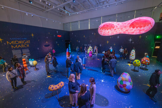 Art installation titled "Midnight March" by Masako Miki. Colorful, textured sculptures and floating forms fill a cosmic-themed room with visitors exploring.