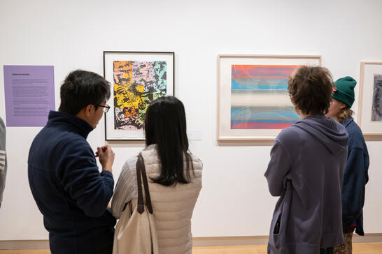 A group of people observes modern art in a gallery, featuring bright abstract paintings. The setting is calm, fostering appreciation and discussion.