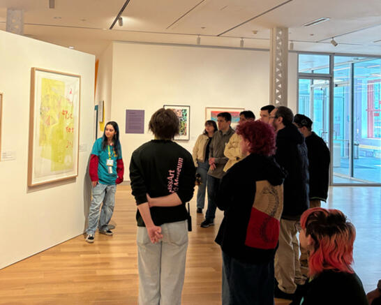 A group of people listens to a guide in front of colorful artworks in a modern gallery. The atmosphere is attentive and curious.