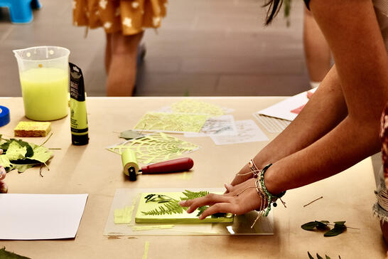 A person arranges leaves on a printing plate at a craft table with art supplies and paper nearby.