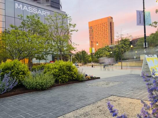 Urban plaza at sunset with MassArt building, trees, flowers, and a tall orange building in the background.