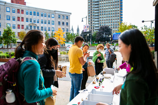 People stand in line at an outdoor table with supplies, smiling and talking, with city buildings in the background.