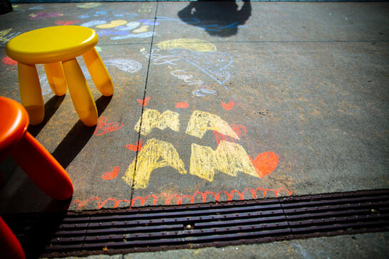 Two small colorful stools beside chalk drawings and writing on a sunlit sidewalk, including I am and red hearts.