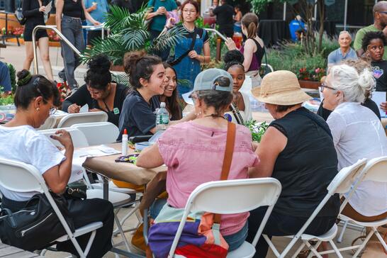 A group of people sit around a table outdoors, talking and crafting at a community event.