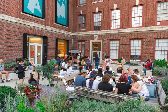 People gather at outdoor tables in a courtyard surrounded by a brick building for a social or creative event.