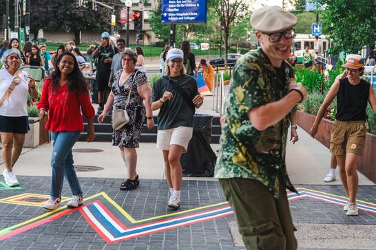 A group of people dance and smile outdoors on a city sidewalk decorated with colorful tape lines.