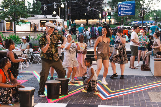 People of all ages dance and play drums outdoors at a lively community event with colorful tape on the ground.