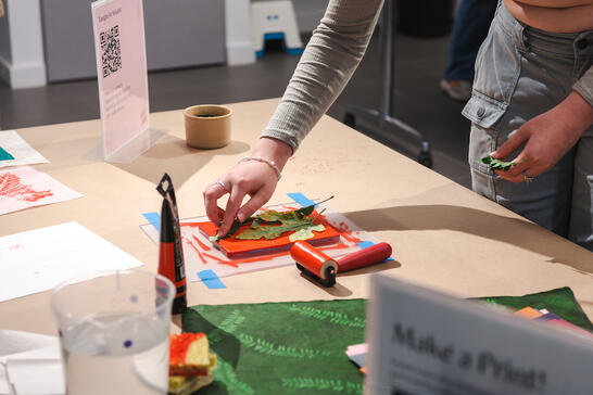 A person uses leaves and paint to make prints on paper at a craft table with art supplies.
