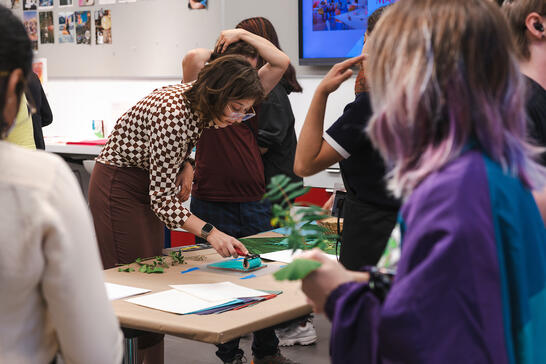 A woman demonstrates art techniques at a table while several people observe and participate in a classroom setting.