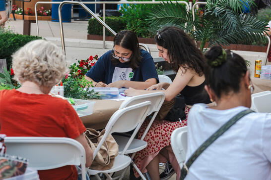 Several people sit at outdoor tables, drawing or crafting together, surrounded by plants and flowers.