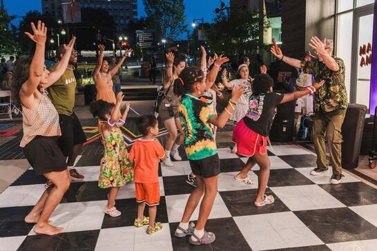 A group of adults and children dance energetically together outdoors on a black and white checkered floor at night.