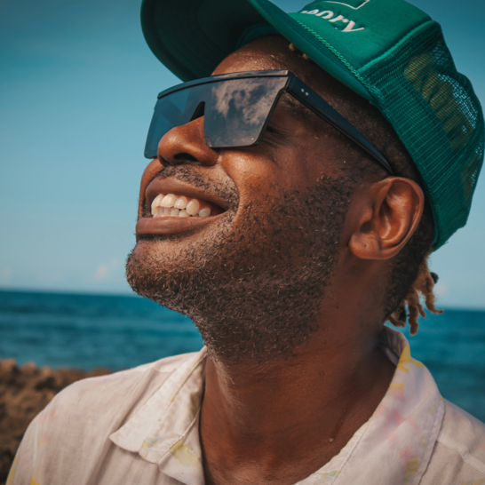 Smiling man in sunglasses and a green cap stands outdoors by the ocean under a blue sky.