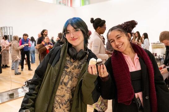 Two smiling people at an indoor event, one holding cupcakes, with a lively group in the background.