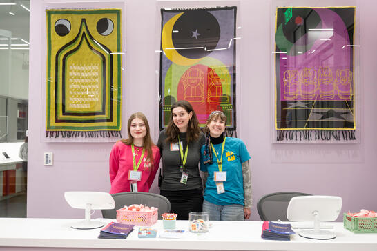 Three women stand smiling behind a desk, with colorful art hanging on the wall behind them.