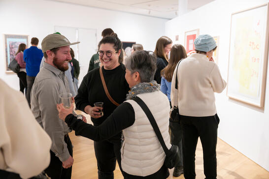 People chatting and smiling at an art gallery, with framed artworks on white walls in the background.