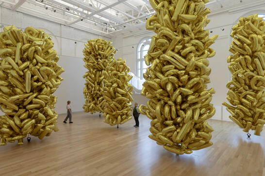 People walk among tall clusters of gold balloons in a bright, spacious gallery with wood floors.