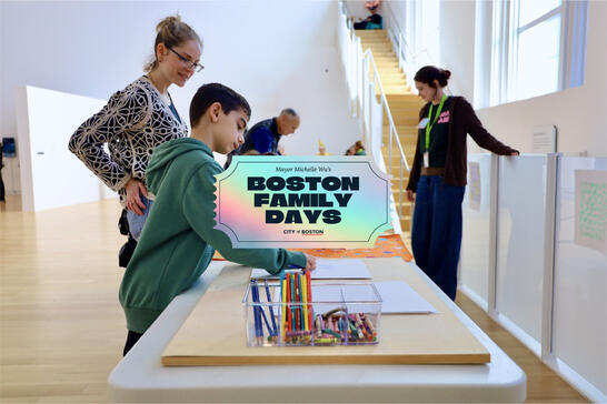 A boy drawing with crayons at a Boston Family Days event, with adults nearby in a bright art space.