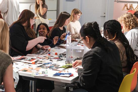 People sit around a table covered with craft supplies, talking and making art projects together indoors.