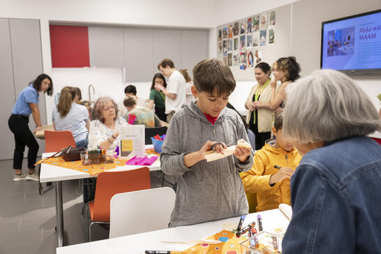 Children and adults work on art and crafts at tables in a bright, busy classroom or activity room.