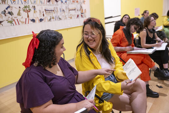 Two people smiling and sharing sketches in an art class, with others drawing in the background.
