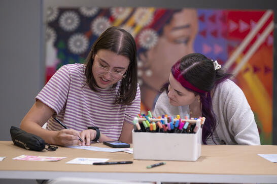 Two young women sit at a table writing and drawing with markers.