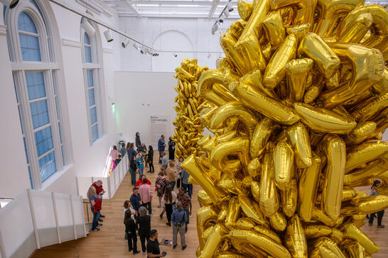 People viewing a large installation of stacked gold foil balloons in a bright museum gallery.