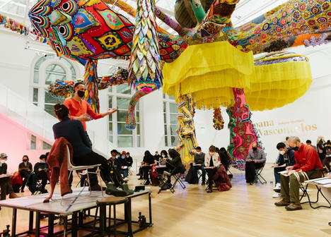 Visitors in the Paine Gallery sitting under a large colorful hanging sculpture drawing from two live models posed on a stage.