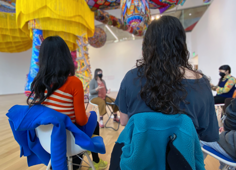 Visitors sit in a circle under a colorful sculpture and installation.