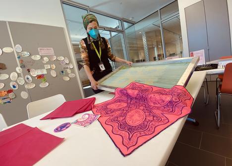 An artist holds a silkscreen with brightly colored and decorated bandanas in on a table