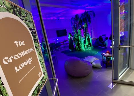 An open doorway with a sign that reads "The Greenhouse Lounge" leading into a room lit with deep purple and green lights and has low seating 