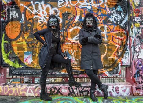 Two women dress in all black with guerrilla masks stand in front of a wall covered with grafitti 